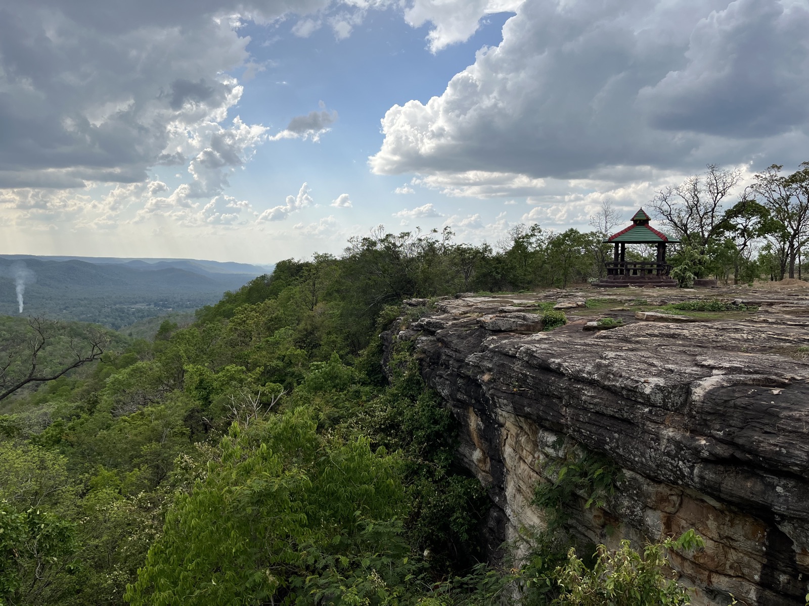 A forested cliff overlooking a wide valley with a small green-roofed pavilion on the rock edge.